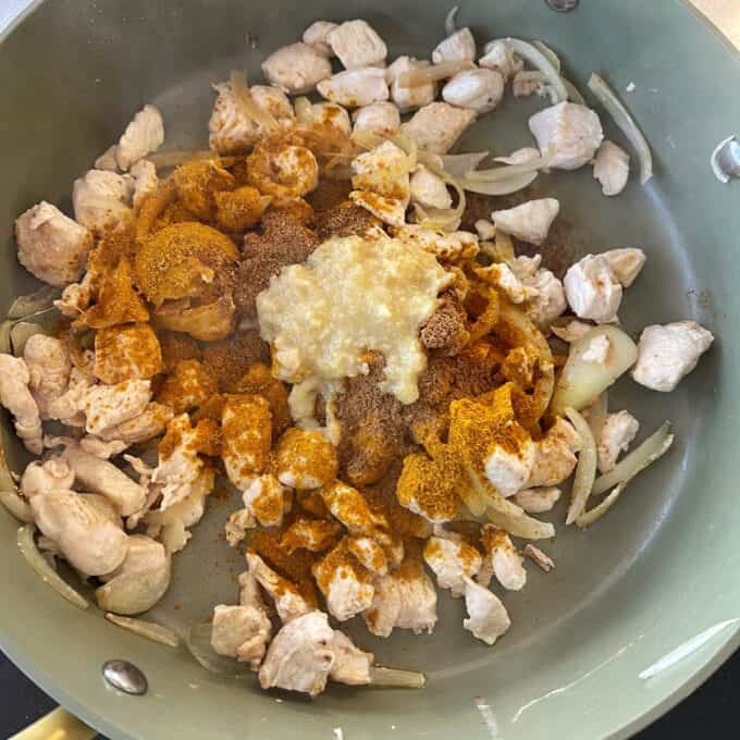 Chicken and Pea Curry Diced chicken and sliced onions cooking in a pan with various ground spices and a mound of minced garlic or ginger added on top.
