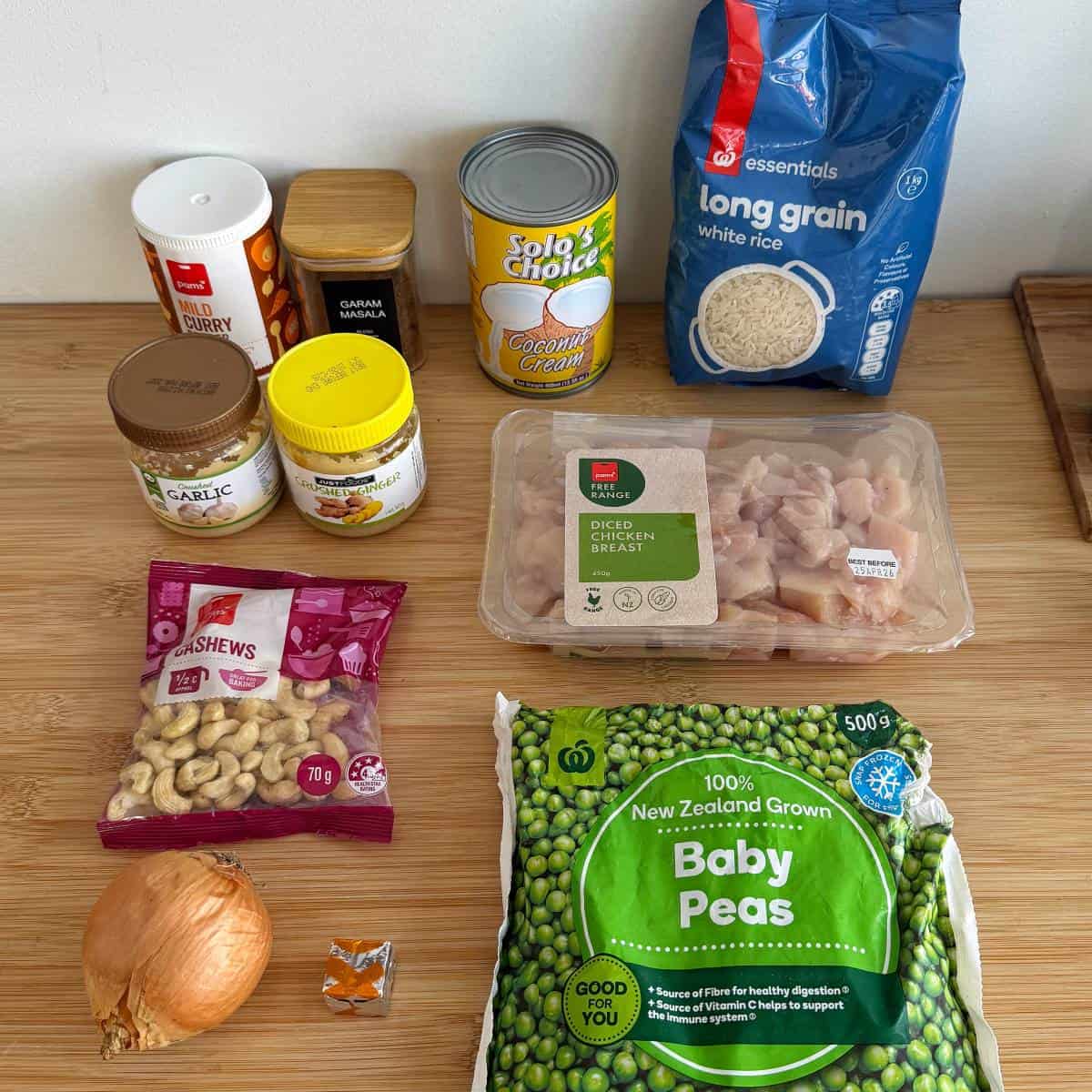 Chicken and Pea Curry Ingredients for a meal arranged on a wooden surface, including diced chicken, rice, baby peas, coconut cream, cashews, spices, onion, garlic paste, stock cube, and coconut oil.
