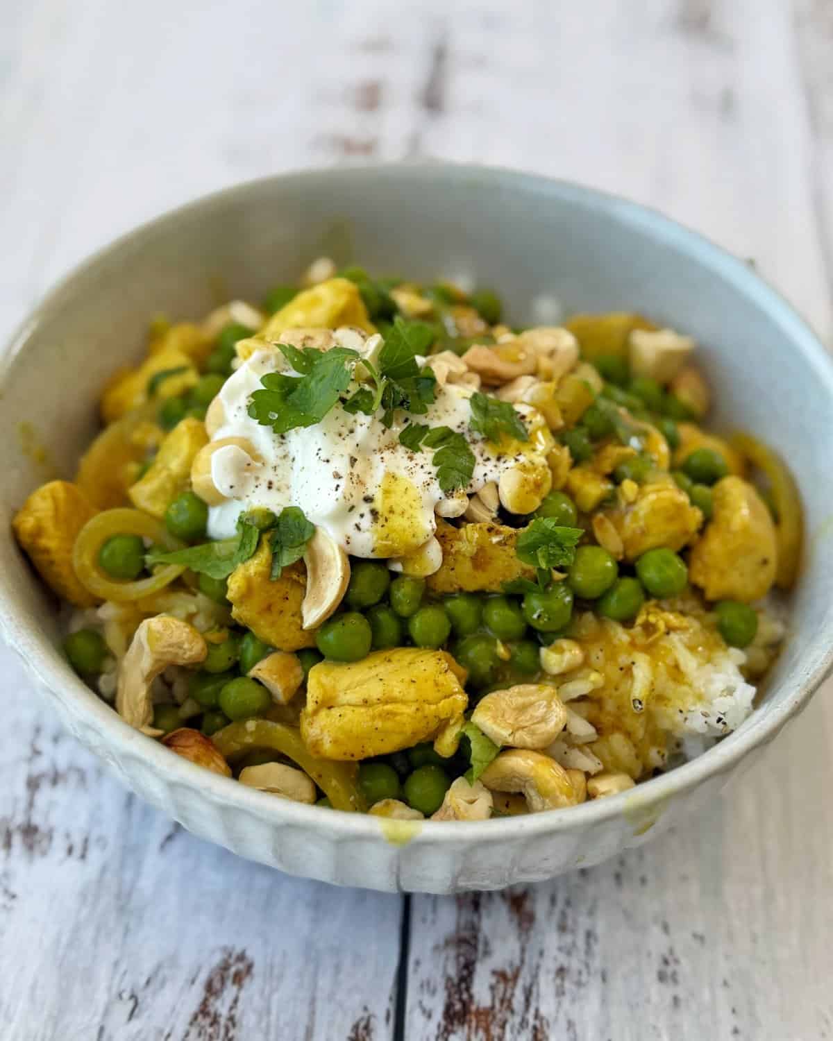 Chicken and Pea Curry A bowl of rice topped with curried chicken, green peas, cashews, a dollop of yogurt, and fresh cilantro on a rustic white surface.