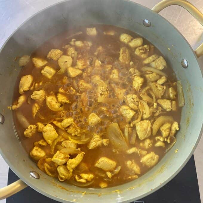 Chicken and Pea Curry A pot of yellow curry with pieces of chicken and sliced onions simmering in sauce on a stove.