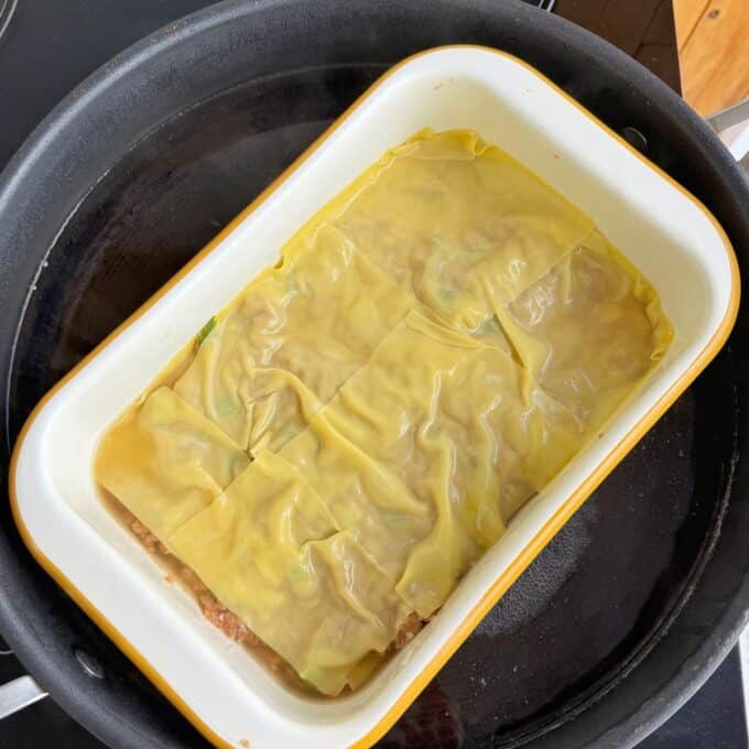 Pork Mince Dumpling Lasagne A rectangular baking dish with uncooked lasagna sheets layered over meat filling, placed inside a larger pan on a stovetop.