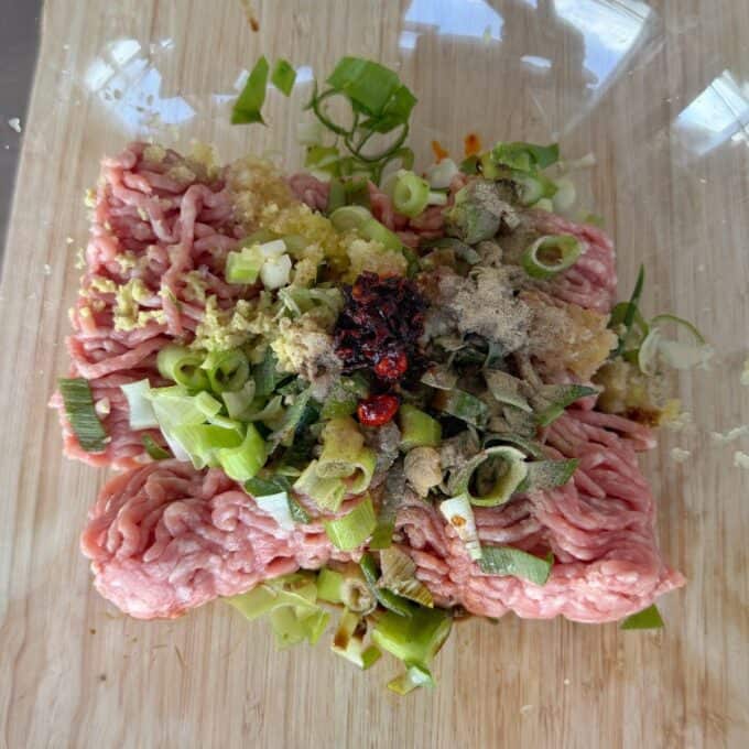 Pork Mince Dumpling Lasagne A pile of raw ground meat topped with chopped green onions, minced garlic, ginger, black pepper, and chili paste on a wooden cutting board.