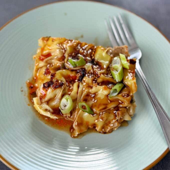 Pork Mince Dumpling Lasagne A serving of lasagna topped with sauce, sliced green onions, and sesame seeds on a green plate with a fork.