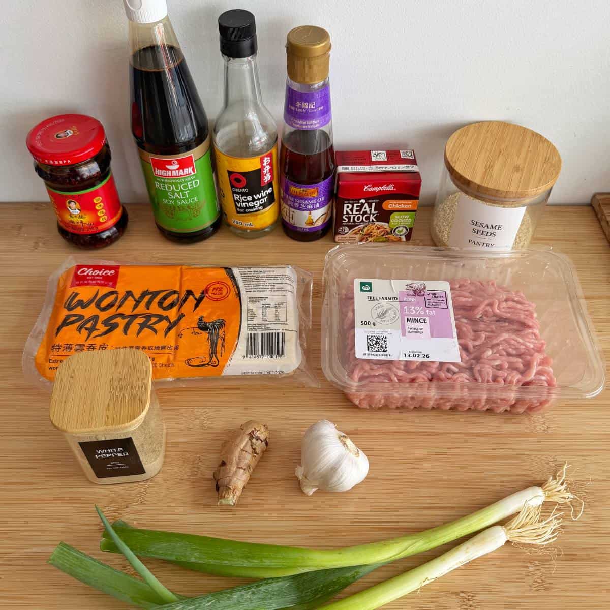 Various ingredients for cooking, including sauces, wonton pastry, pork mince, garlic, ginger, spring onions, sesame seeds, and seasonings, arranged on a wooden surface.