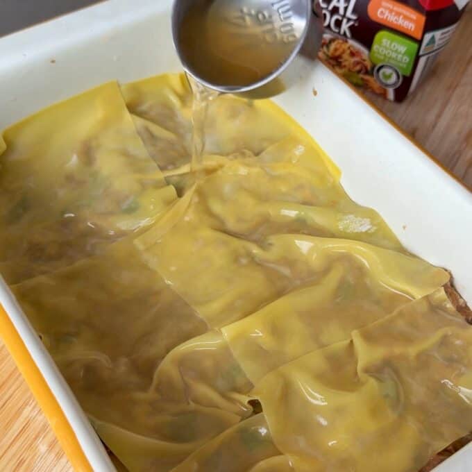 Pork Mince Dumpling Lasagne A hand pours broth over a layer of uncooked lasagna sheets in a white baking dish on a wooden surface.