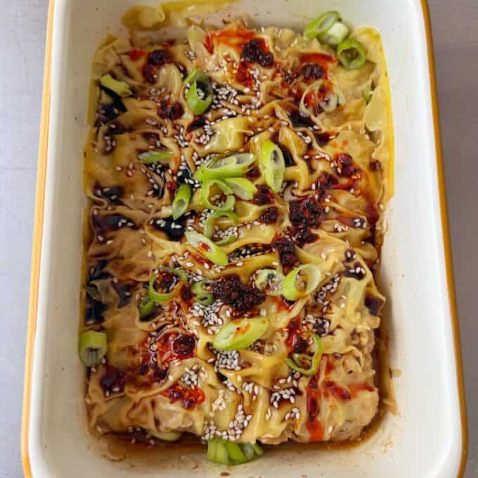 Pork Mince Dumpling Lasagne A baked dish of dumplings topped with sliced green onions, sesame seeds, and a drizzle of chili oil in a rectangular ceramic dish.
