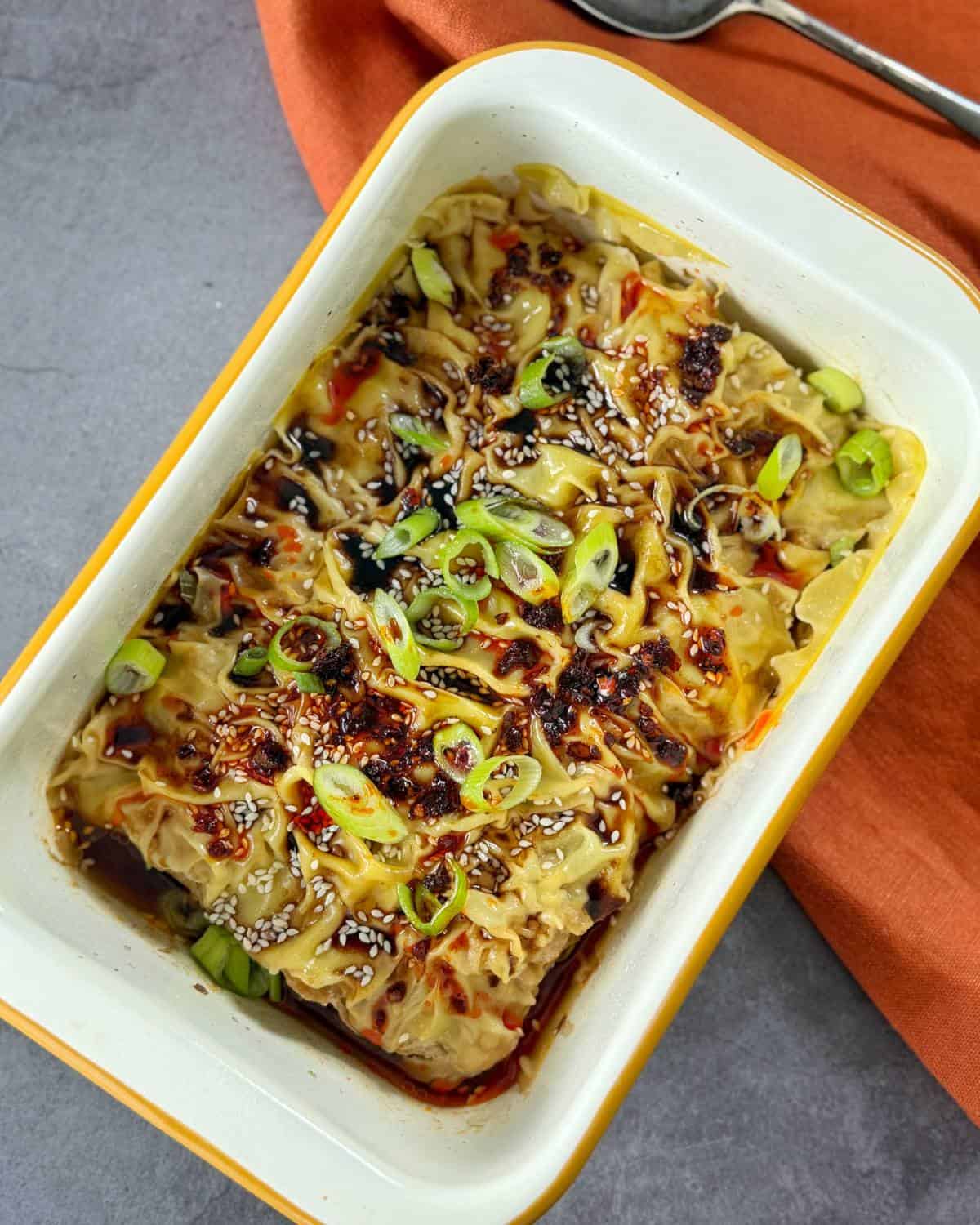 Rectangular dish with dumplings topped with chopped green onions, sesame seeds, and chili oil sauce on a gray surface with an orange napkin.