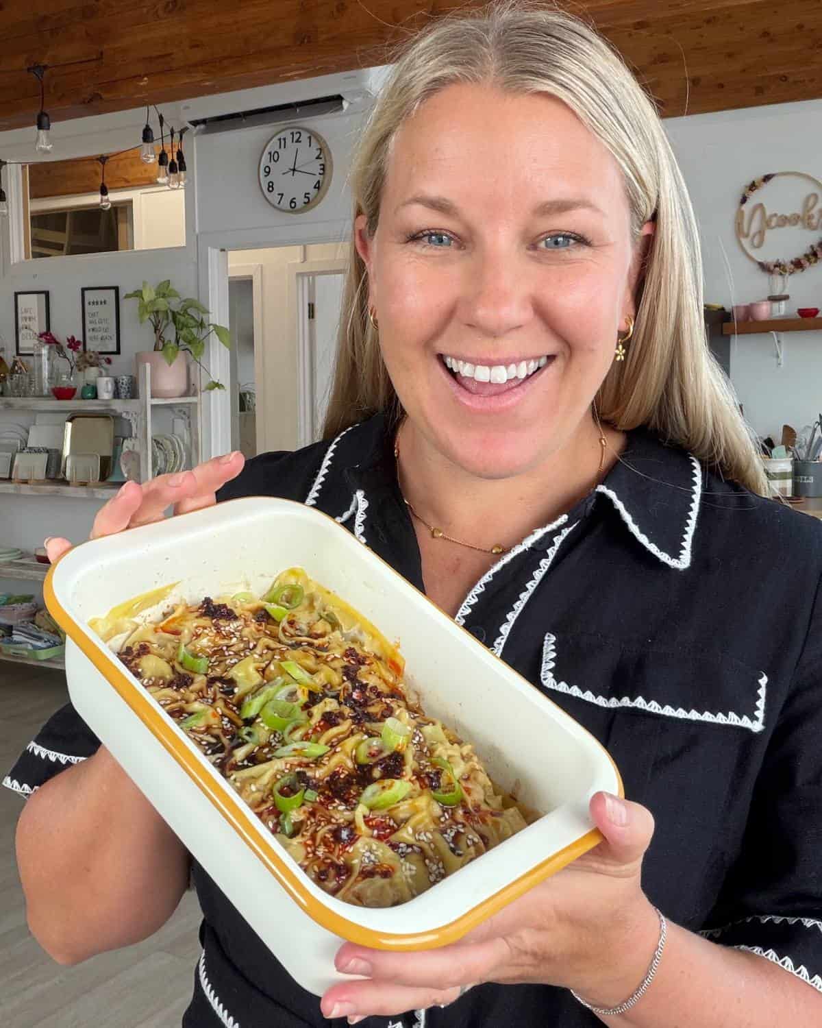 Pork Mince Dumpling Lasagne A woman in a black shirt holds a rectangular dish of noodles topped with sliced green onions and sauce, smiling at the camera in a bright kitchen.