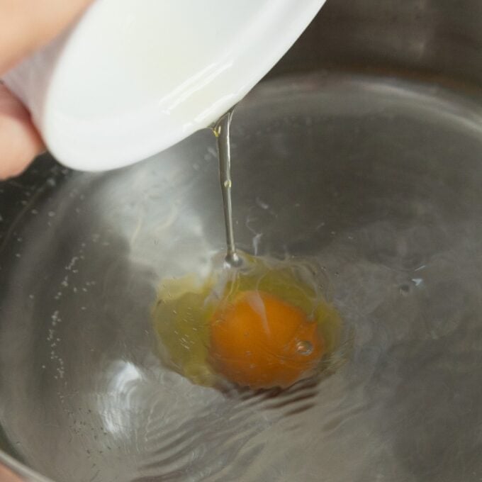 Eggs Benedict with Hollandaise A raw egg is being poured from a small white bowl into a pot of hot water.