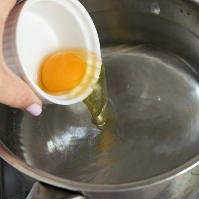 Eggs Benedict with Hollandaise A hand pours a raw egg from a small white bowl into a pot of hot water, preparing to poach the egg.