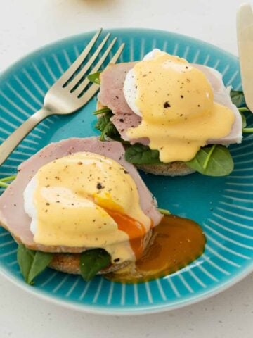 Two eggs Benedict on English muffins with ham, spinach, and hollandaise sauce, served on a turquoise plate with a fork and knife. One egg has a runny yolk.