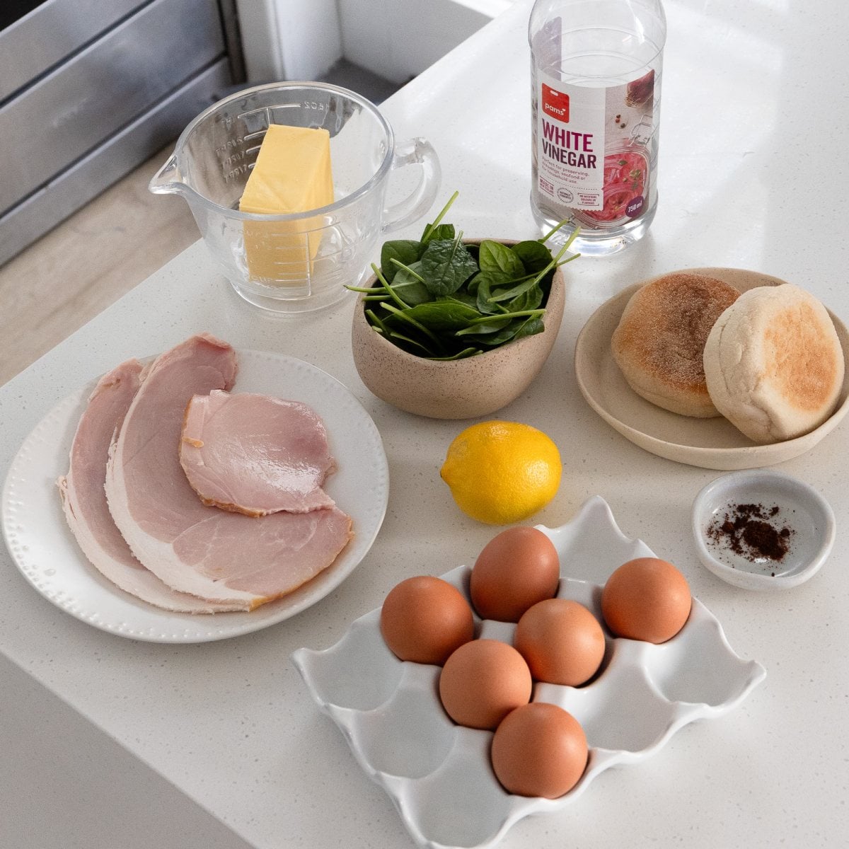 Eggs Benedict with Hollandaise Ingredients for eggs Benedict arranged on a countertop: eggs, ham, butter, spinach, lemon, white vinegar, English muffins, and ground coffee in a small dish.