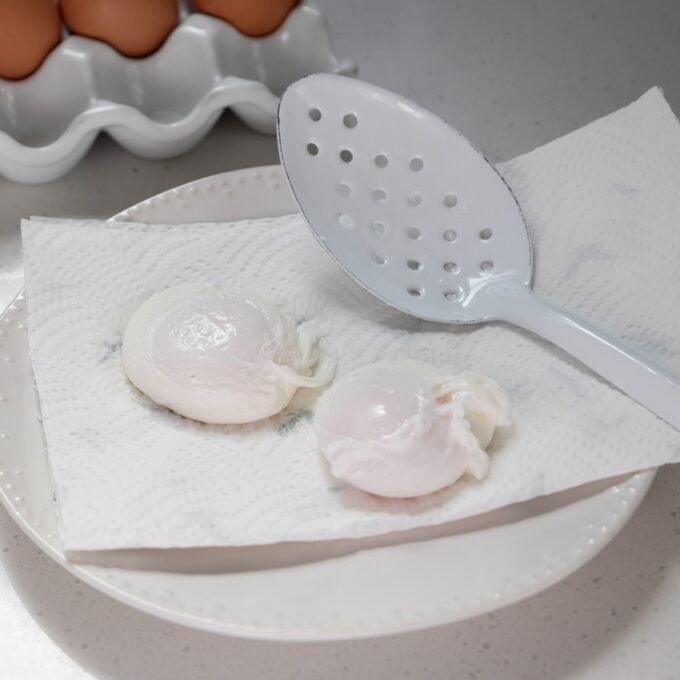 Eggs Benedict with Hollandaise Two poached eggs rest on a paper towel atop a white plate, with a slotted spoon beside them and a carton of eggs in the background.