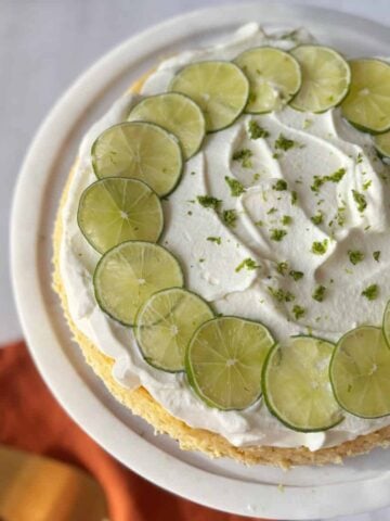 A round cake topped with whipped cream, lime slices arranged in a circle, and lime zest, displayed on a white cake stand.