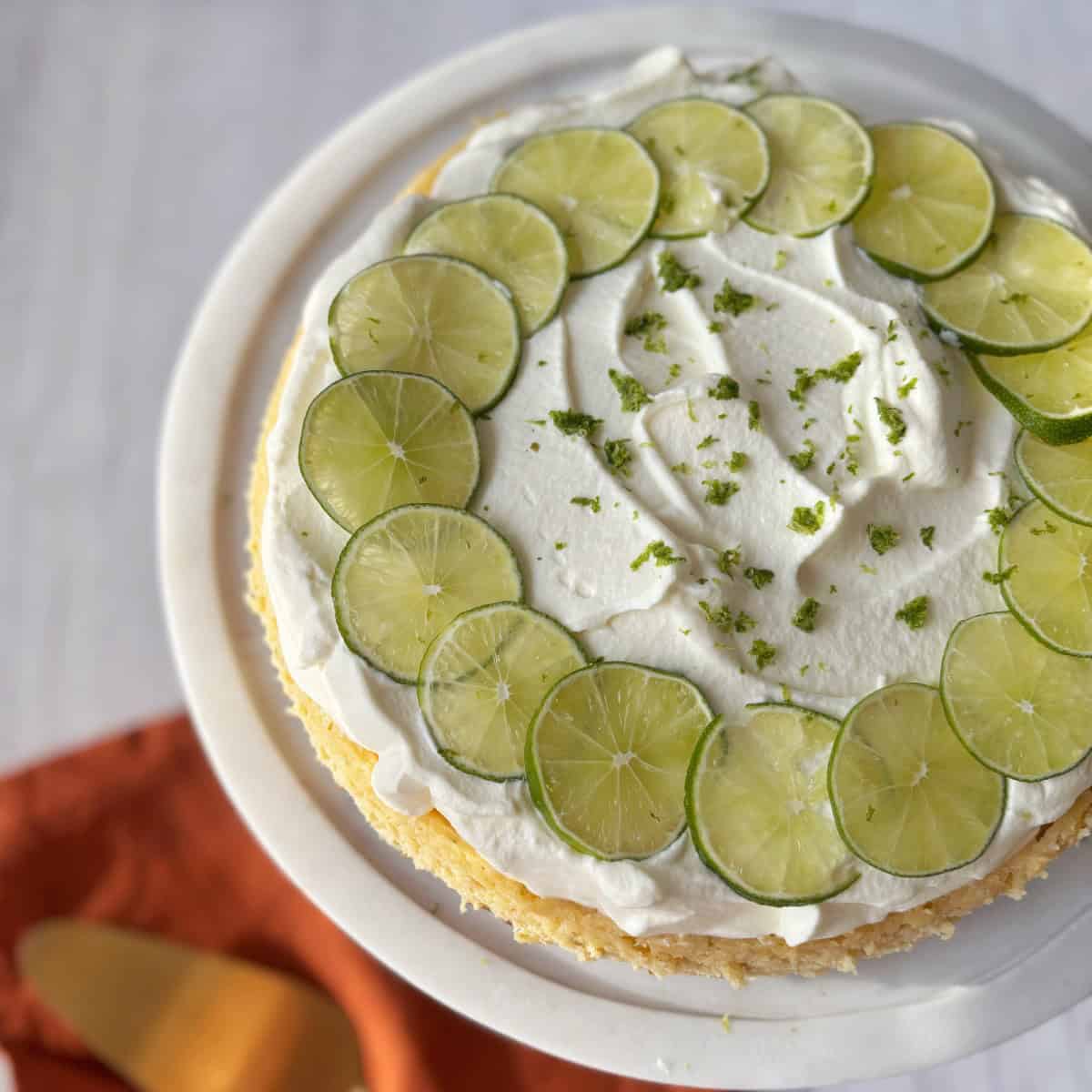 Lime Gingernut Cheesecake A round cake topped with whipped cream, lime slices arranged in a circle, and lime zest, displayed on a white cake stand.