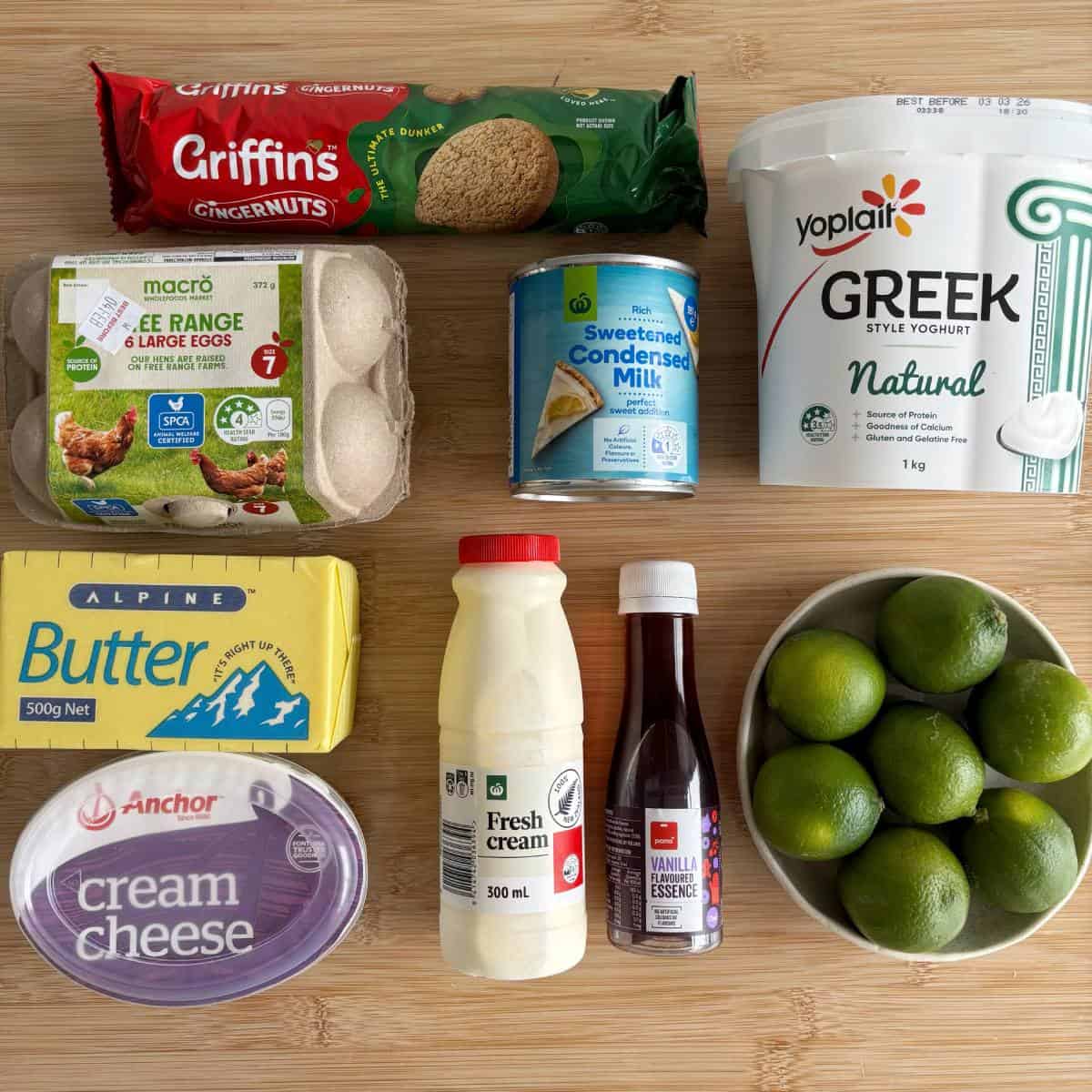A selection of ingredients on a table: eggs, gingernut biscuits, sweetened condensed milk, Greek yogurt, butter, fresh cream, vanilla essence, cream cheese, and a bowl of limes.
