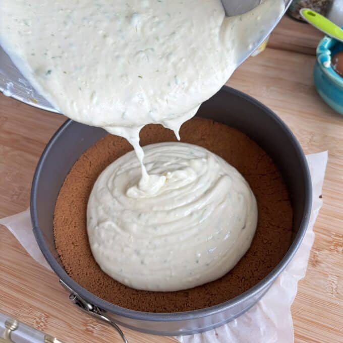 Lime Gingernut Cheesecake Thick cheesecake batter being poured into a springform pan with a graham cracker crust on a wooden surface.