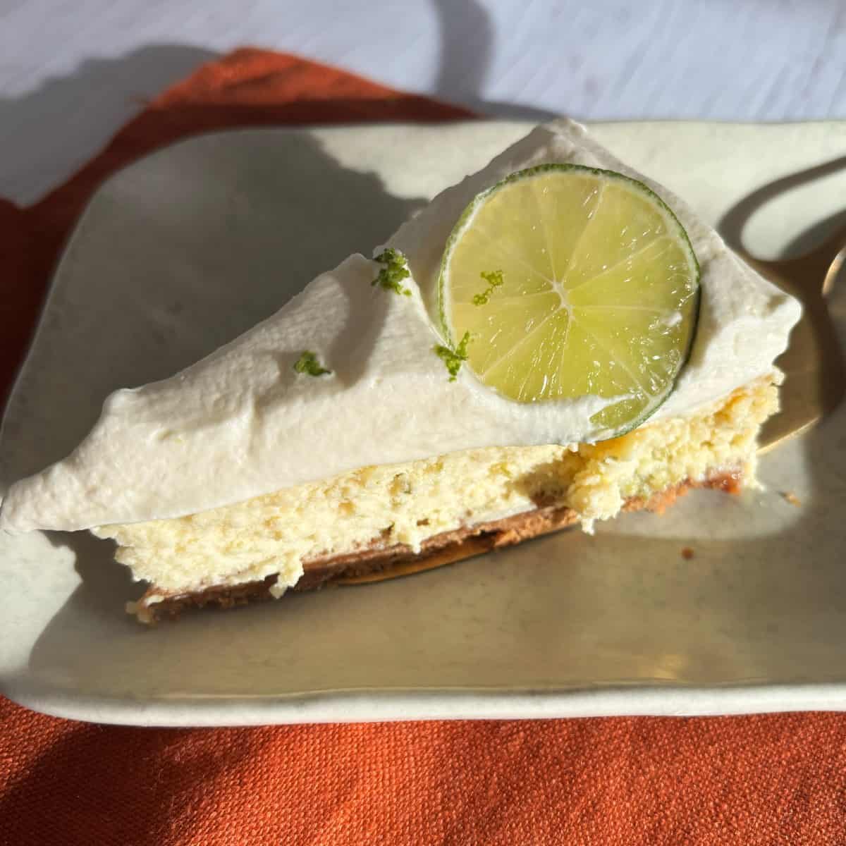 Lime Gingernut Cheesecake A slice of key lime pie with whipped topping and a lime slice garnish on a square plate, set on an orange napkin.