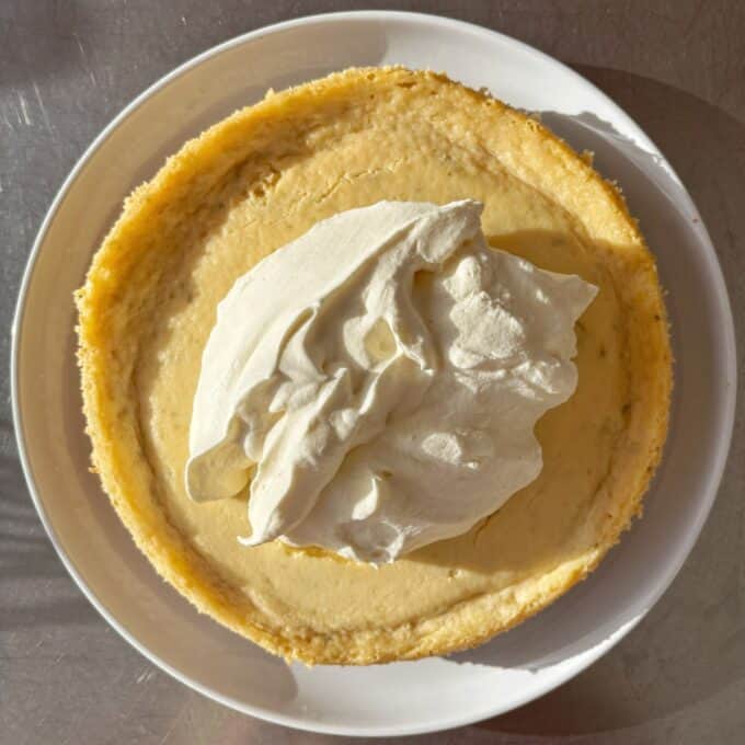 Lime Gingernut Cheesecake A plain cheesecake on a white plate topped with a large dollop of whipped cream, photographed from above.