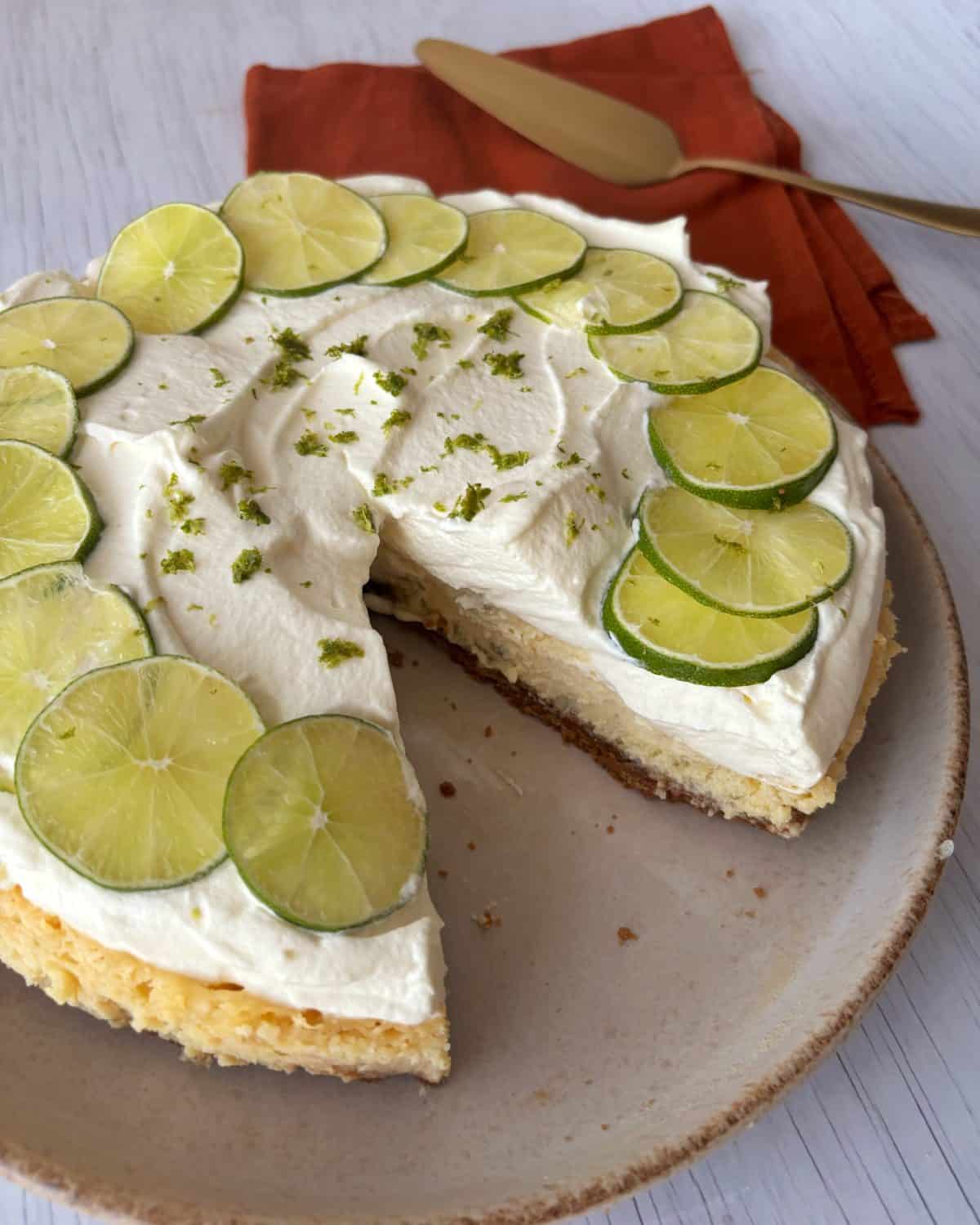 A key lime pie with a slice missing, topped with whipped cream, lime slices, and lime zest, sits on a beige plate next to a gold cake server and an orange napkin.