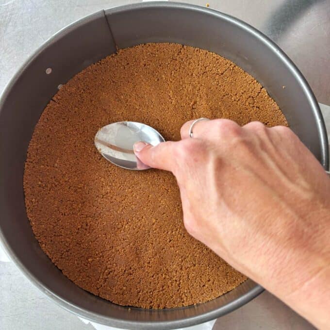 Lime Gingernut Cheesecake A hand uses a spoon to press down a crumb crust in a round metal baking pan on a countertop.