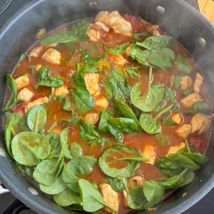 Marry Me Chicken Penne Pasta A pot of simmering stew with chunks of chicken, fresh spinach leaves, and tomato-based sauce.