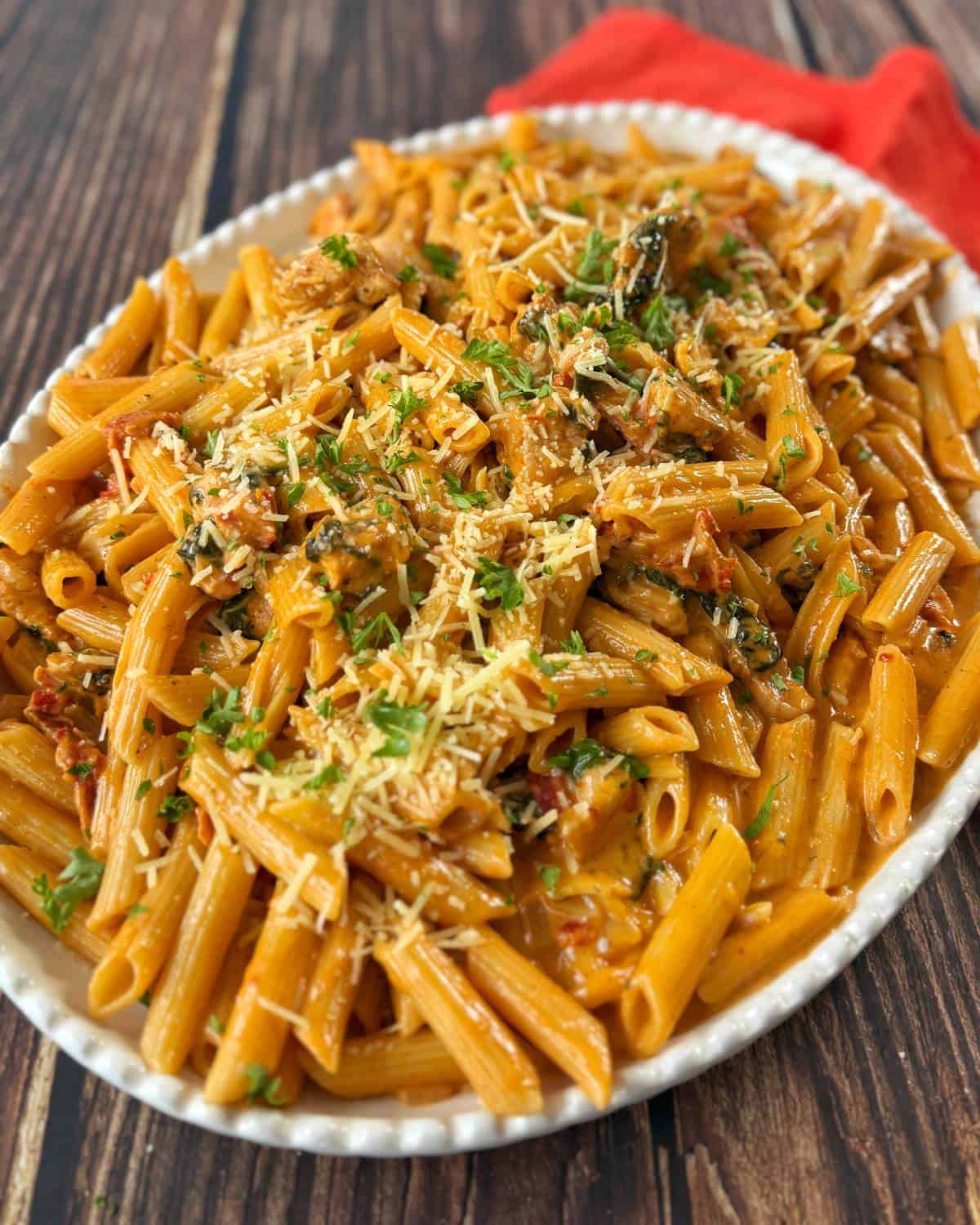 A white oval dish filled with penne pasta in a creamy sauce, topped with grated cheese and chopped fresh herbs, on a wooden surface with a red napkin.