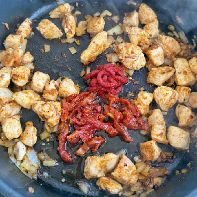 Marry Me Chicken Penne Pasta Chicken pieces, diced onions, sun-dried tomatoes, and tomato paste cooking in a black skillet.