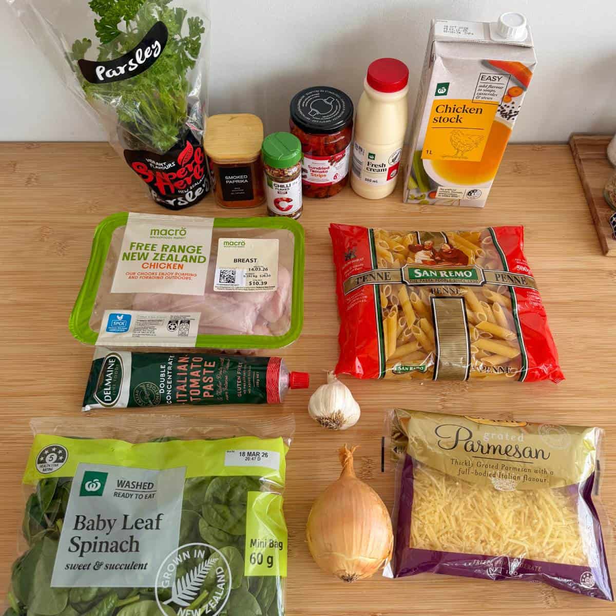 Ingredients are arranged on a wooden surface, including chicken breast, pasta, spinach, onion, garlic, tomato paste, chicken stock, parmesan, parsley, and various seasonings.