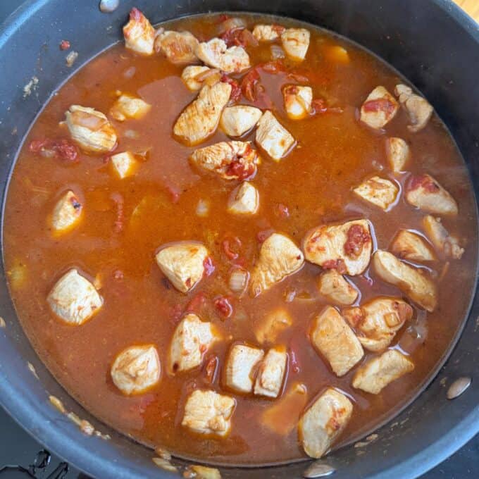 Marry Me Chicken Penne Pasta Chunks of chicken simmer in a reddish-brown sauce with visible tomato and onion pieces in a black pan.