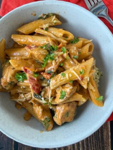 A bowl of penne pasta with creamy sauce, chicken pieces, sun-dried tomatoes, and herbs, served on a wooden table with a red napkin and fork.