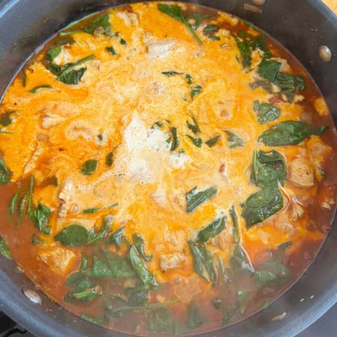 Marry Me Chicken Penne Pasta A pot of orange-colored soup or stew with pieces of meat and green leafy vegetables simmering in broth.