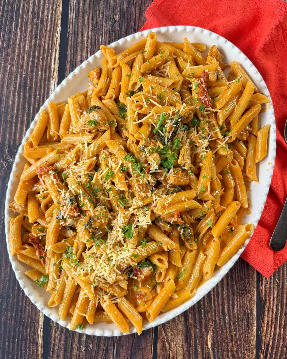 Marry Me Chicken Penne Pasta An oval white dish filled with penne pasta in a red sauce, topped with shredded cheese and chopped herbs, on a wooden table with a red napkin.