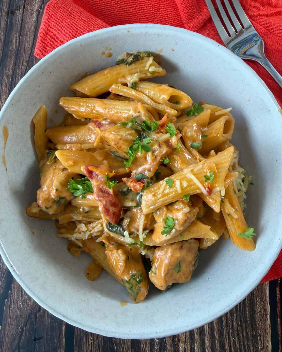 Marry Me Chicken Penne Pasta A bowl of penne pasta with creamy sauce, grilled chicken pieces, sun-dried tomatoes, spinach, and parsley, garnished with grated cheese. A fork and red napkin are beside the bowl.