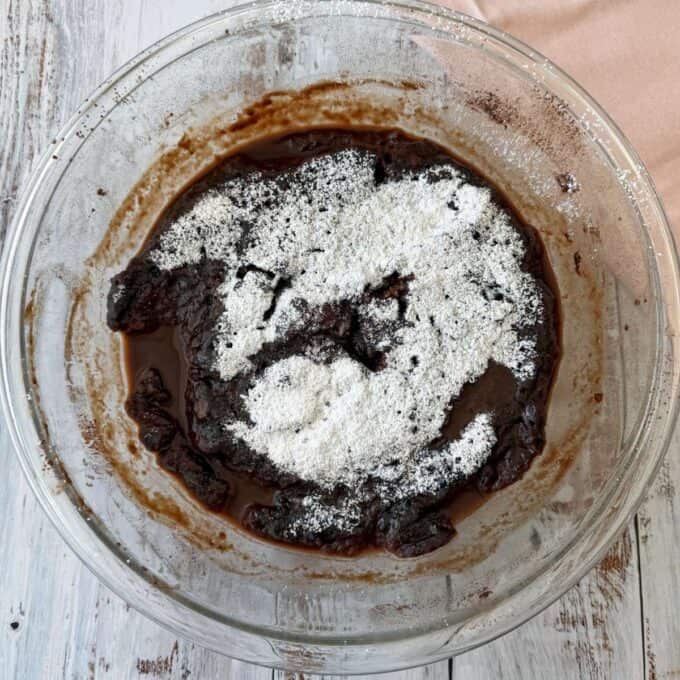 Microwave Self-Saucing Chocolate Pudding A glass bowl containing chocolate batter with a layer of white flour on top, placed on a rustic white wooden surface.