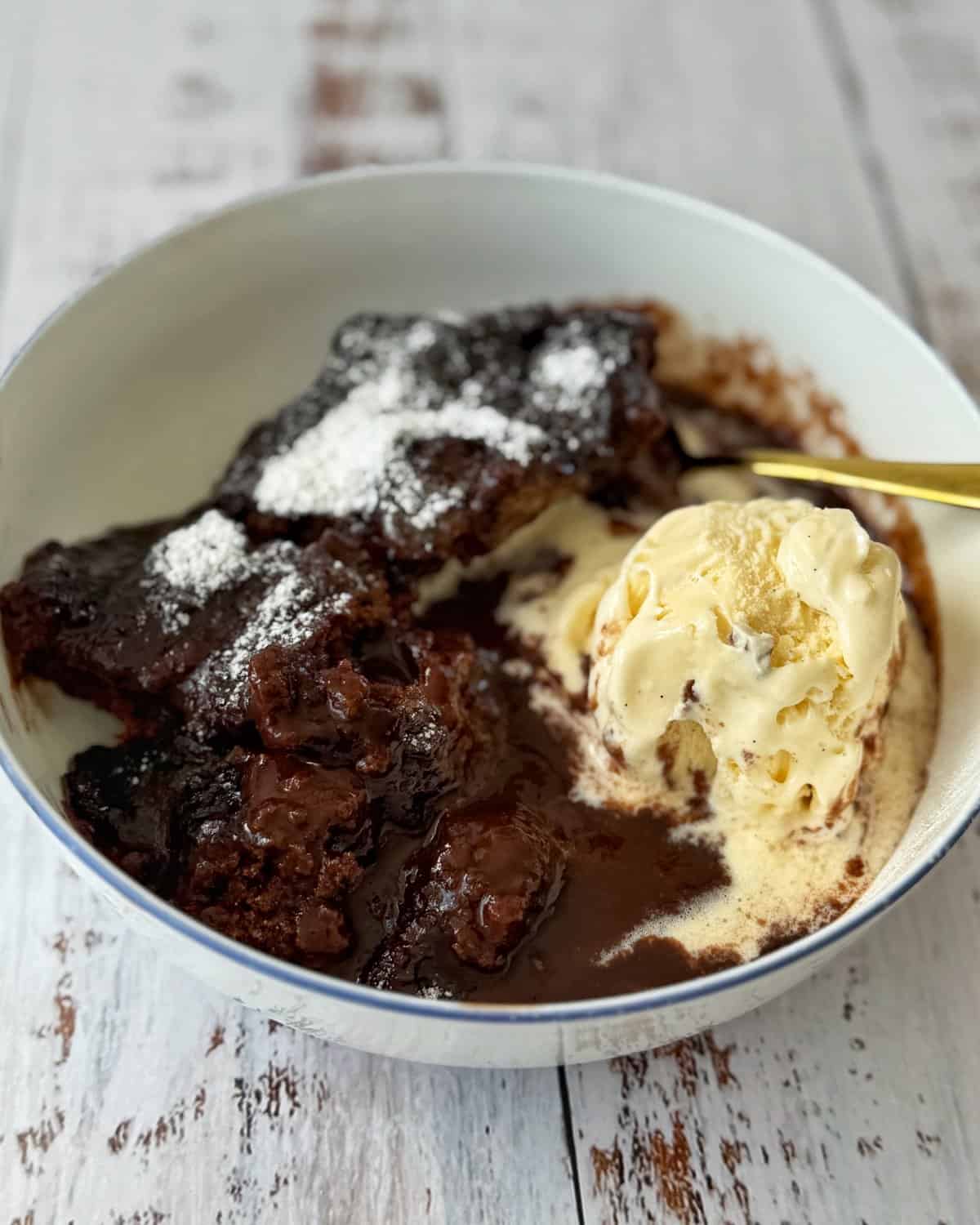 Microwave Self-Saucing Chocolate Pudding A bowl of chocolate lava cake topped with powdered sugar, served with a scoop of vanilla ice cream on a rustic white wooden surface.