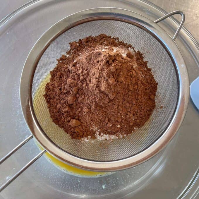 Microwave Self-Saucing Chocolate Pudding A metal sieve containing cocoa powder and flour is placed over a glass bowl with melted butter underneath.