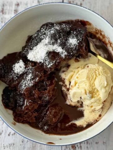 A bowl of chocolate pudding cake topped with powdered sugar, served with a scoop of vanilla ice cream, on a light wooden surface.