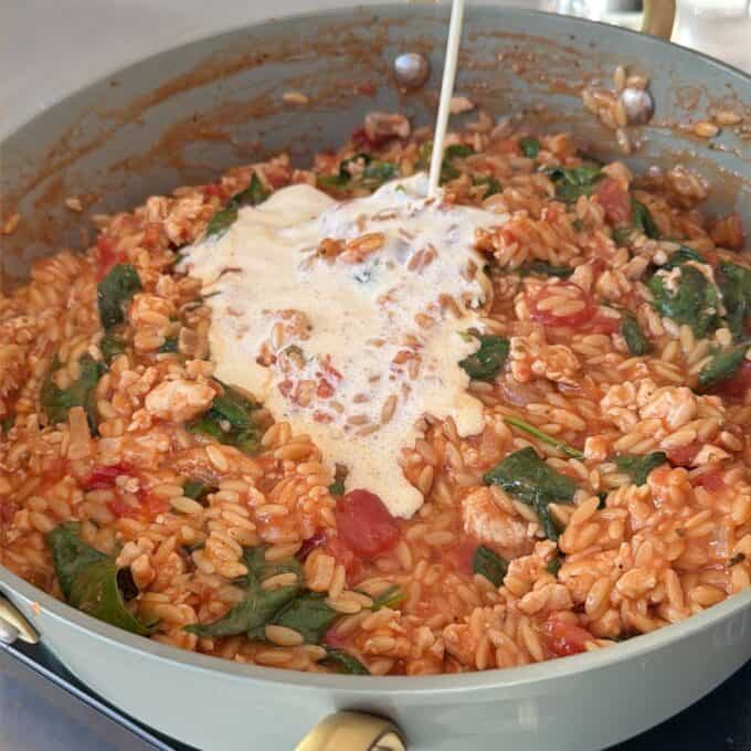 One Pot Creamy Chicken Orzo A skillet of cooked orzo with spinach, tomatoes, and chicken, with cream being poured over the mixture.