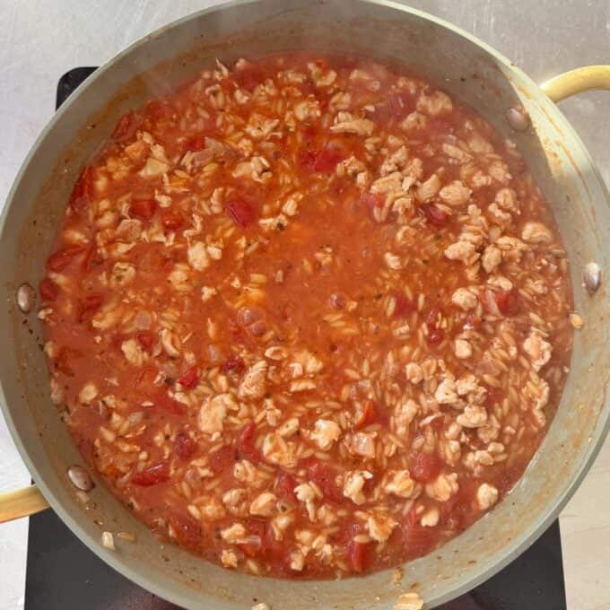 One Pot Creamy Chicken Orzo A pan filled with a simmering mixture of ground meat, rice, diced tomatoes, and sauce on a stovetop.