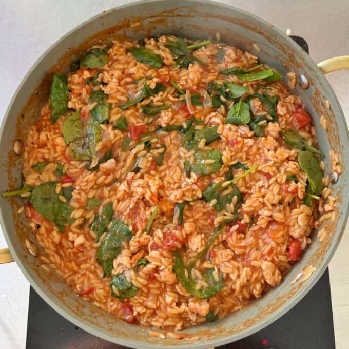 One Pot Creamy Chicken Orzo A pan filled with cooked orzo, spinach, chopped tomatoes, and pieces of chicken in a tomato-based sauce.