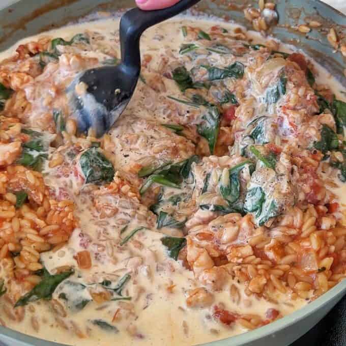 One Pot Creamy Chicken Orzo A creamy rice dish with spinach, tomatoes, and chicken is being stirred in a pan with a black spatula.