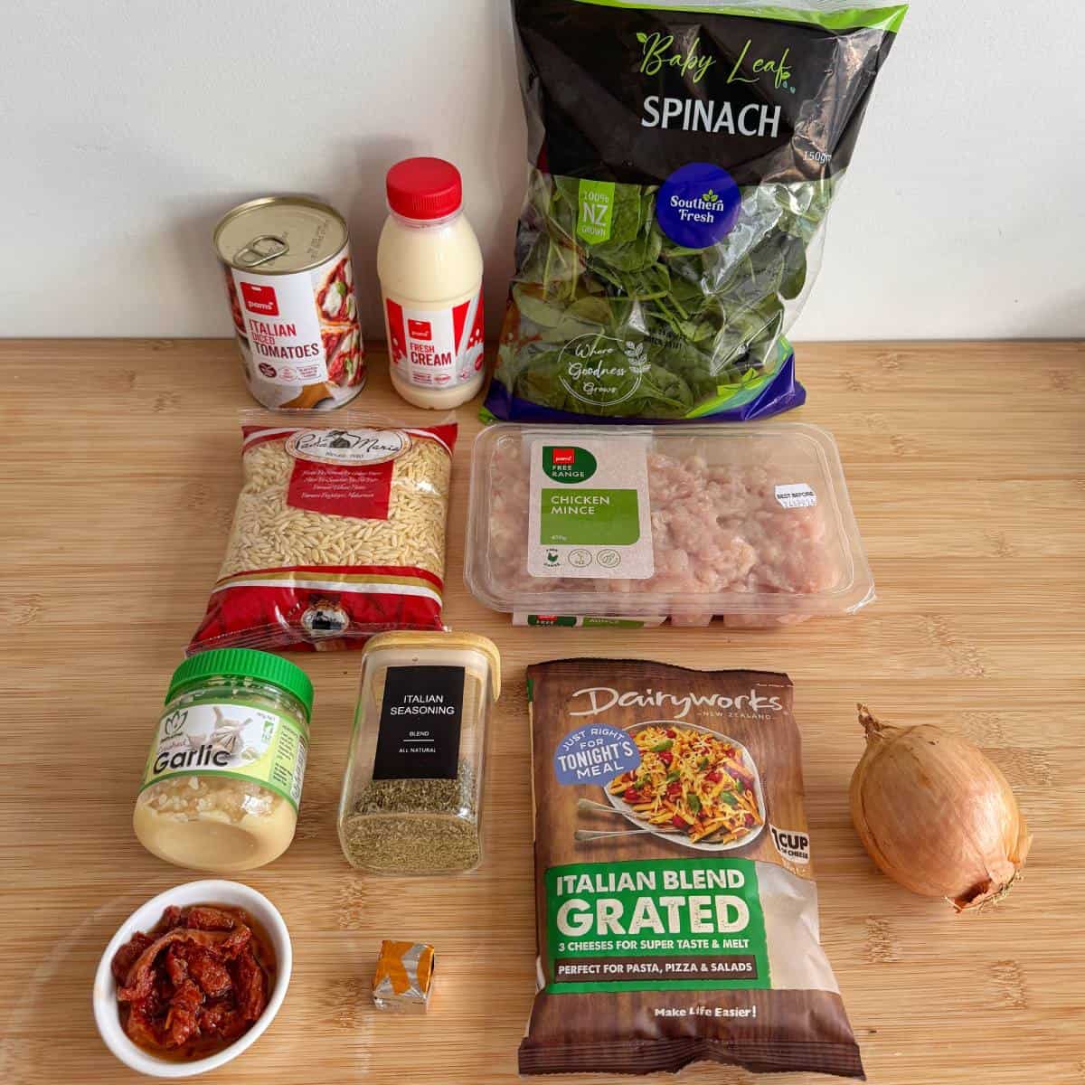 One Pot Creamy Chicken Orzo Ingredients for a recipe on a wooden surface, including spinach, tomato cans, cream, chicken mince, rice, garlic, Italian seasoning, cheese, onion, a stock cube, and sun-dried tomatoes.