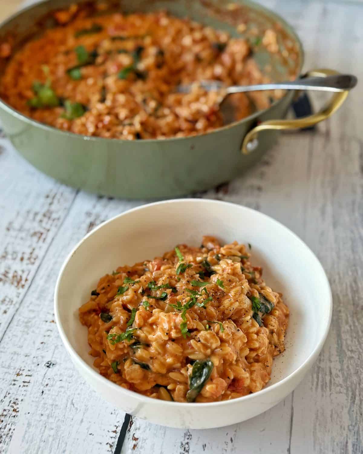 One Pot Creamy Chicken Orzo A bowl of creamy tomato risotto garnished with herbs sits on a white table, with a pan of risotto and a serving spoon in the background.