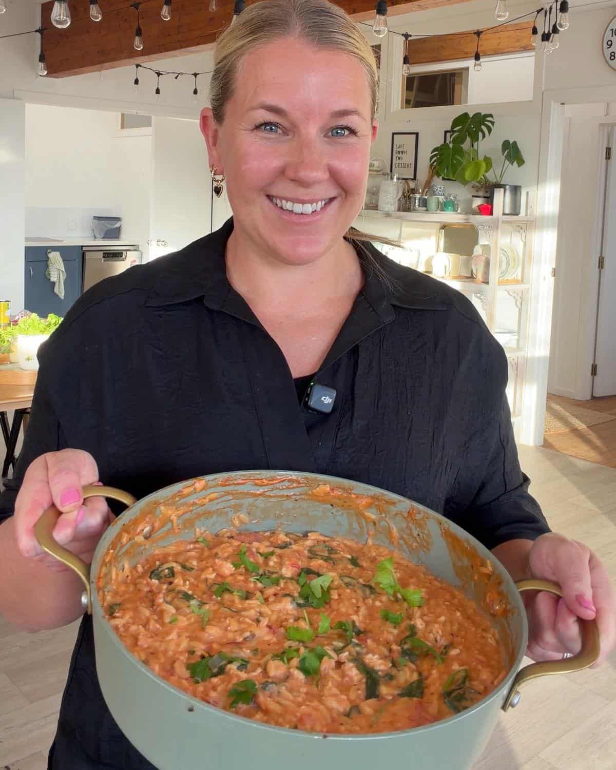 One Pot Creamy Chicken Orzo A woman in a black shirt smiles while holding a large pot filled with a creamy tomato-based rice dish topped with chopped herbs in a bright, modern kitchen.