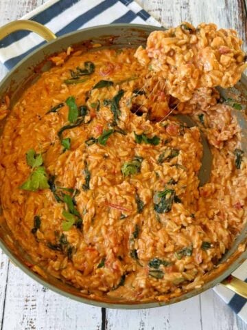 A pan of creamy tomato and spinach risotto sits on a wooden table, with a spoon lifting a portion and melted cheese stretching from the pan.