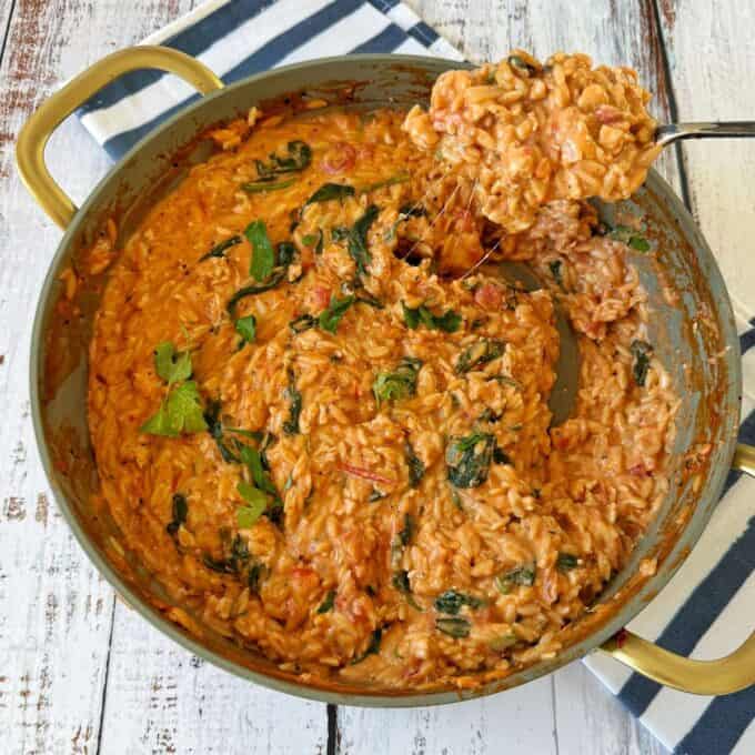 One Pot Creamy Chicken Orzo A pan of creamy tomato and spinach risotto sits on a wooden table, with a spoon lifting a portion and melted cheese stretching from the pan.