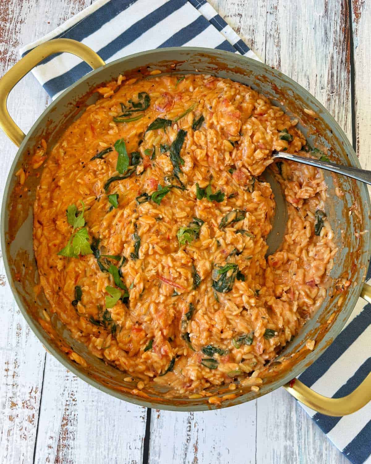 One Pot Creamy Chicken Orzo A large pot of creamy tomato and spinach risotto sits on a striped cloth, with a spoon resting inside the pot.