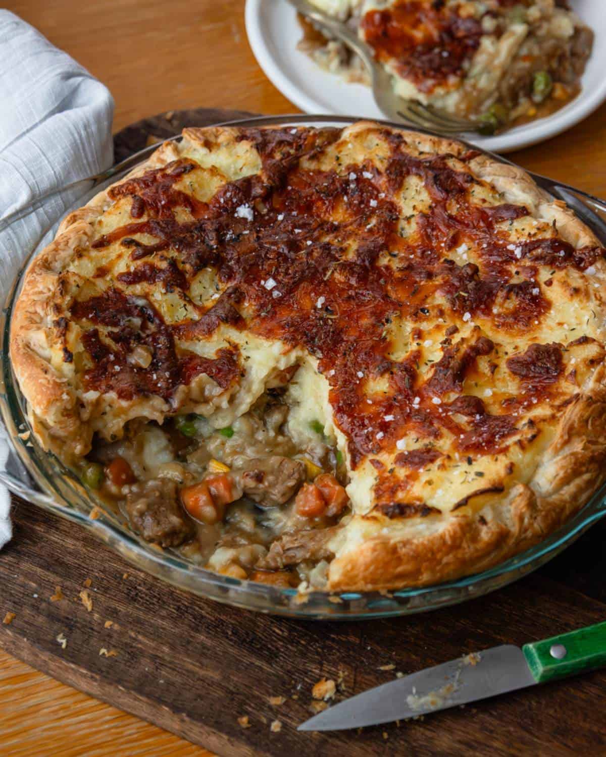 Shepherd's Pie A baked meat and vegetable pot pie with a slice removed, showing the filling, sits on a wooden board with a knife beside it; a plate with a serving is in the background.