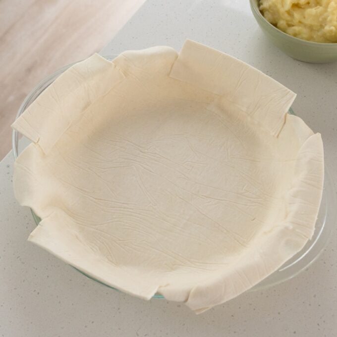 Shepherd's Pie A round glass pie dish lined with a sheet of uncooked pastry dough, with excess edges hanging over the sides. A bowl of mashed potatoes is visible in the background.
